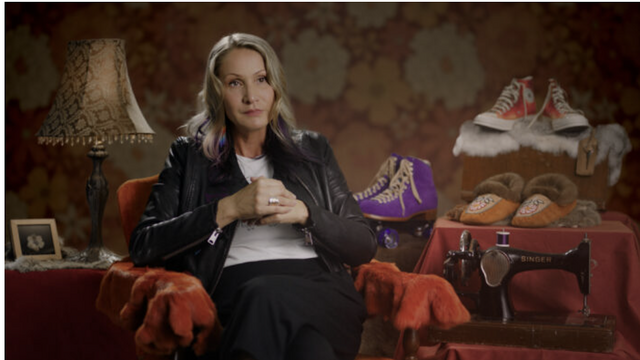 A woman sits in a chair. She is surrounded by homey features, a lamp with a beaded shade, a photo on a table. Three pairs of footwear are featured, a pair of red high top sneakers, a pair of purple suede lace up ankle boots, and a pair of beaded moccasins with fur around the tops.