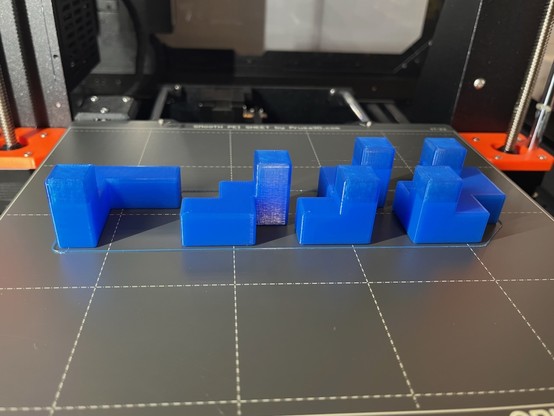 A set of puzzle pieces, rather like if Tetris pieces bent in three dimensions, on a 3D printer bed. The top third-ish is *almost* the same color of blue plastic as the rest of them…