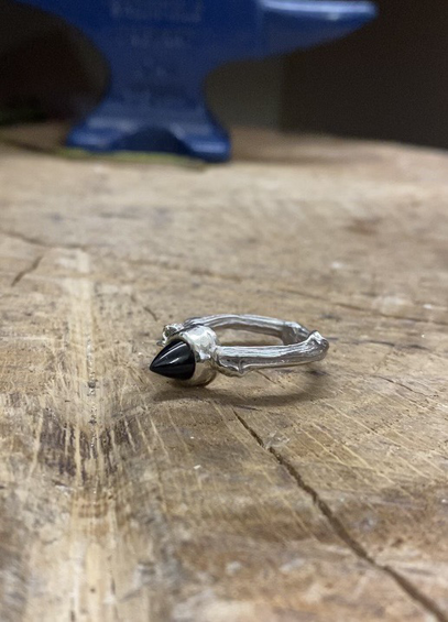 A silver ring lies on its side on a block of wood. The shank of the ring is uneven and organic, the stone is a pointy bullet shaped black onyx.