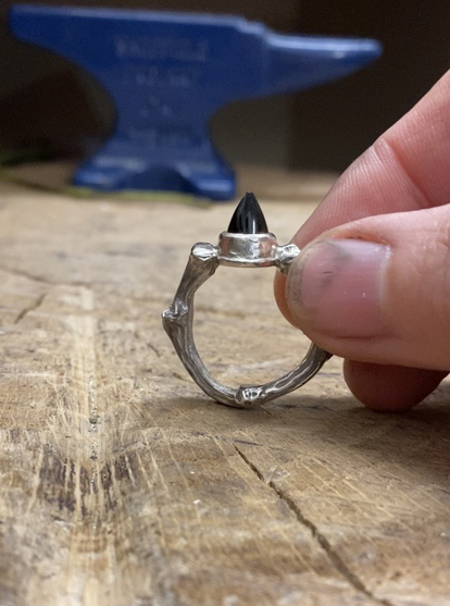 A hand with dirty nails holds a ring  on a block of wood. The shank of the ring is uneven and organic, the stone is a pointy bullet shaped black onyx.