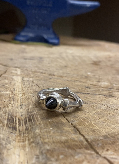 A silver ring lies on its side on a block of wood. The shank of the ring is uneven and organic, the stone is a pointy bullet shaped black onyx.