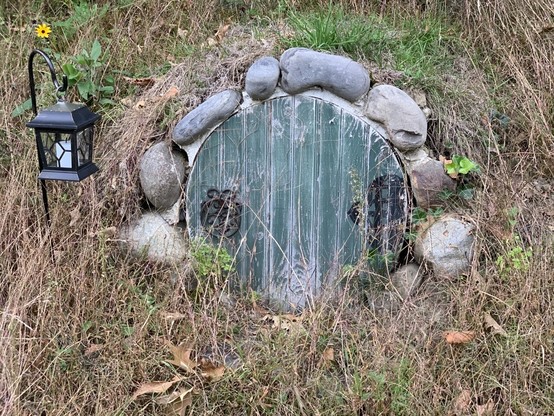 A round, green, wooden door with a black metal knocker is built into the side of a hill. A lantern hangs to the left of the door on a metal post.