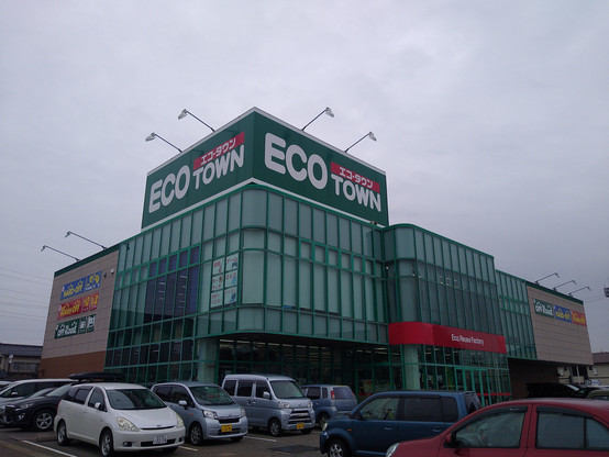 Exterior photo of a green-window paned building with a large banner at the top indicating the building name, "ECO-TOWN". Photo taken in a car parking lot.