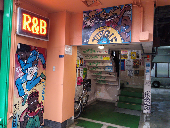 Exterior stairs leading up to Record Jungle. Groupings of graffiti including a man holding a saxophone jumping over a woman, with a large R&B sign above it. The Record Jungle logo is a grouping of folks playing drums with happy expressions. Various band posters and advertisements can be seen on the stairs leading up. The exterior facade is painted a light orange, and the flooring is a darker green.