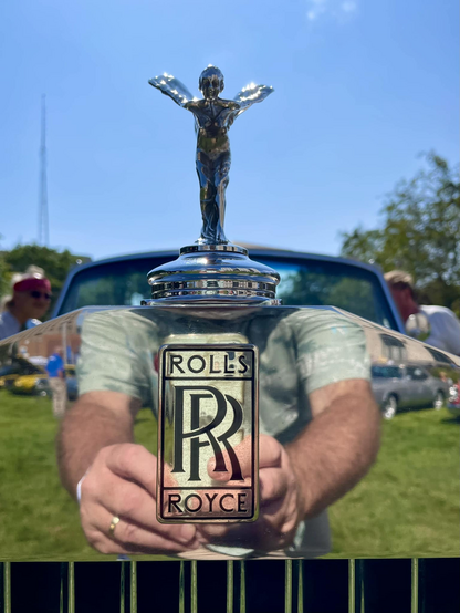 A Rolls-Royce "Spirit of Ecstasy" hood ornament on the top of a reflective strip atop the chrome grill and badge with a blue sky in the background. The photographer is reflected in the reflective top of the grill so that his cell-phone camera is where the badge is and we can see his arms reaching out with his shoulders at the top of the grill with the hood ornament taking the place of his head.