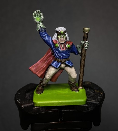 Heroquest Wizard miniatures. Classic pose with a staff and hand raised casting a spell. They have a red cape, a blue tunic, and cream trousers. Their eyes and hand and the gold on their staff all have a green glow.
