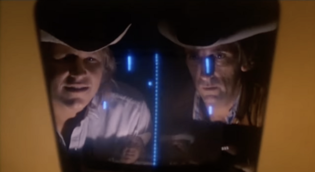 Image from the film "Rancho Deluxe" showing Harry Dean Stanton playing a Pong arcade machine. His face is reflected in the screen and he is wearing a cowboy hat.