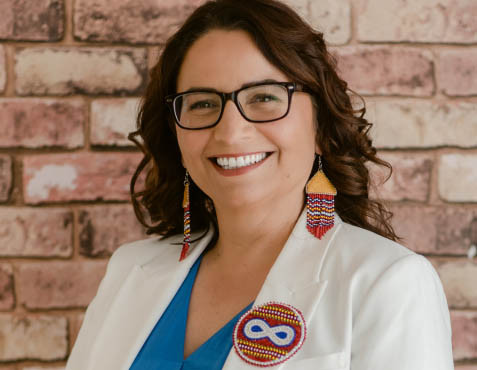 A woman smiles widely toward the camera. She wears a white jacket and a beaded pin with a Métis infinity symbol on it.