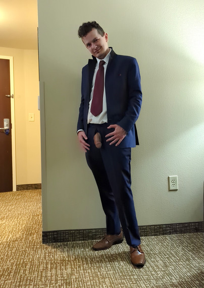 Photo of a guy wearing a full navy blue tuxedo standing in front of a pale wall, looking at the camera. His suit jacket is open showing his white dress shirt and maroon red tie, and he's wearing brown dress shoes. The fly of his pants is undone and his flaccid penis and balls are fully hanging out of his pants.