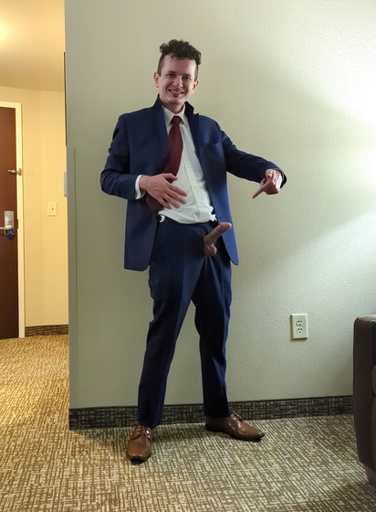 In this photo his dick is fully erect and is pointed upwards at a diagonal, 45 degree angle. His left hand is out in front of him, pointing a finger back at his hard dick and balls which are fully exposed outside of his pants, while the whole rest of his body is dressed up in his tuxedo, and he is grinning at the camera.
