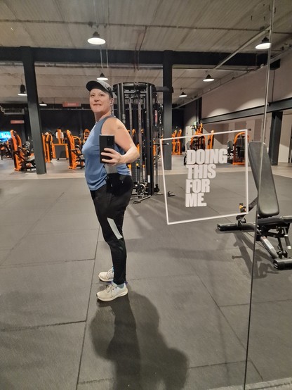 Me, standing in front of the mirror. 1,70 meters, 85,7 kilo's. Wearing a black cap, blue/purple sleeveless shirt, black pants and white shoes. On the mirror a sticker with the text "Doing this for me". Next to me a bench and in the background more fitness machines.