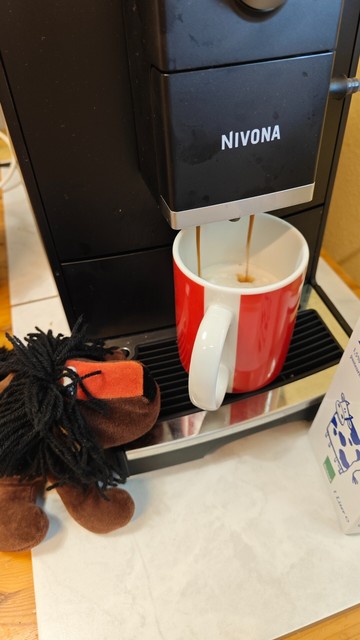 Kaffee-Vollautomat produziert Espresso in den Cappucino in eine rote ver.di Tasse, daneben Perlsacklöwe Afrolöwe