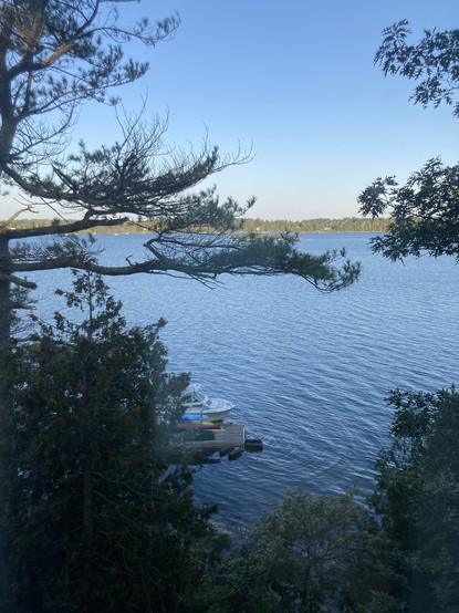 Morning on the coast of Maine. Blue sky, blue water seen through some pine trees. The bow of a small motor boat is tied up at a dock. The end of some canoes and kayaks are on the dock. Itâ€™s a little crisp temperature-wise.