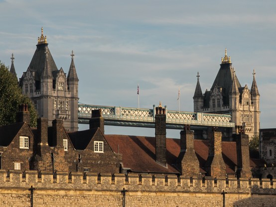 The towers and upper crossing of London's Tower Bridge is visible behind and above houses with red sloping roofs and tall chimneys inside a crenellated stone castle wall.