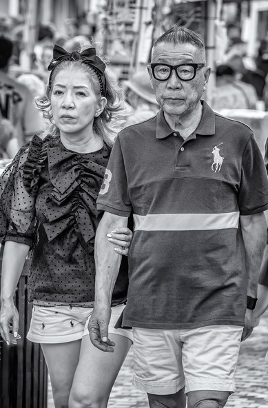Older couple in Polo branded clothing walk down a street in Honolulu. He wears distinctive glasses with a heavy black frame and has his hair spiked into a peak. She wears a hair band with a bow on top and a ruffled blouse.