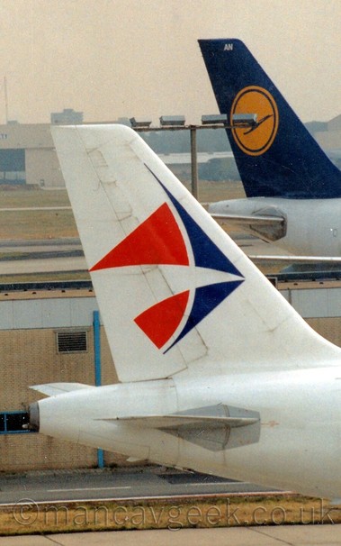 Closeup of the tail of a twin engined jet airliner taxiing from left to right.
The plane is mostly white, with a blue and red logo on the tail that is somewhere between a child's kite, and an angel fish.
In the background, a low, sandy coloured airport building can be seen, with the back end of a jet airlinervwith a white body and a blue and yellow tail just beyond that.
In the far distance, a large white hangar with the doors partially open in the middle, and the words "safety - security" on the upper section, is slowly disappearing into haze, under a hazy grey sky.