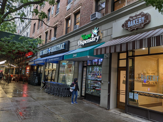 A photo of a NYC street. In the back is a shop called Tal's Bagels, next to the Kosher Marketplace, next to Vape & Dispensary, next to By the Way Bakery (a kosher, gluten free, dairy free, parve bakery)