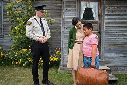 A police officer stands with his hands resting on his belt buckle looking at a mother and son outside of an unpainted wood-framed home. The mom's arm is around the boy and he has a stuffed full brown leather valise clutched in his hand.