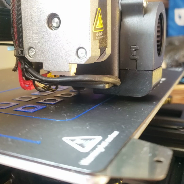 Close video of a 3d printer extruder printing with blue filament. The print is still short, and consists of multiple squares with circular voids in the centre.
