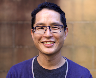 Writer Guan Un, an Asian man with short black hair and glasses. He wears a T-shirt, a purple cardigan, and a dimply smile.