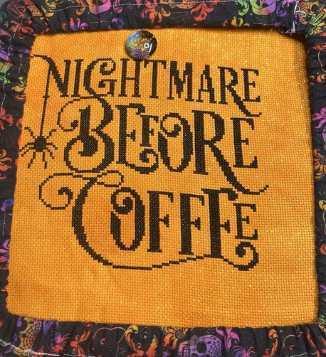 A completed cross stitch on orange opalescent fabric. The words “Nightmare Before Coffee” are stitched in black, and there is a small spider midway down the left side.