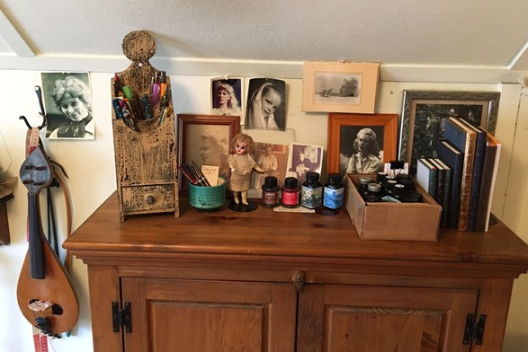 View of a wooden bookcase top where one can see a collection of fountain pens, ink bottles, journals, and old family photos. A tiny antique doll is on a stand near the pens, and a fiddle-like musical instrument hangs on the wall.