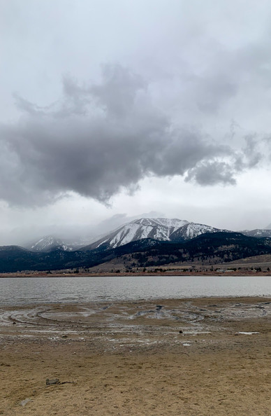 Tall photo of shore of a shallow desert lake, with snow-capped mountains and a cloudy sky in the background
