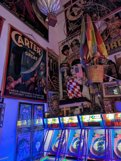 A corner photo of skee-ball machines, the wall above and to the side is covered in colorful posters, many of which appear like movie posters from the mid 1900's. There is a model of a balloon and basket with a doll figure inside, beside a statue of an Elias Brothers Big Boy.
