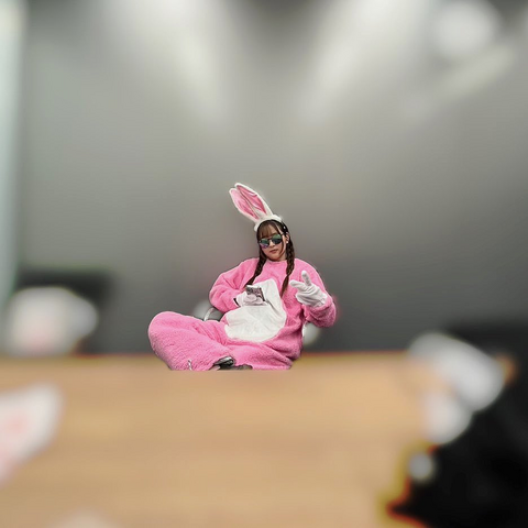 Sally in a pink onesie wearing baddit ears and shades looking at her phone while giving a v sign as most of the picture is blurred out.