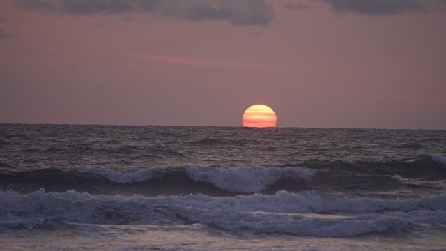 Pink orange sun emerges from the ocean horizon, waves break on sure. A few clouds at the top of the picture, blue against the pink lavender sky.
