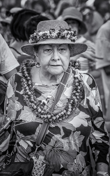 Older woman wearing a hat with a flower lei around the crown walks down a crowded street in Honolulu with a determined look on her face.