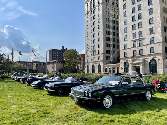 A row of vintage British cars on a green lawn with a vintage brick skyscraper behind. A green 1999 Jaguar XJ Vanden Plas saloon is at the front of the line to the bottom right with its front facing left, with an MGB and several old Jaguars stretching out behind it.