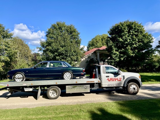 A green 1999 Jaguar XJ Vanden Plas saloon faces left on the back of a flatbed truck that is going to the right on a dirt road. The truck's cab is white with "ARNAZ" on the door. Lawn in the foreground with green deciduous trees behind.