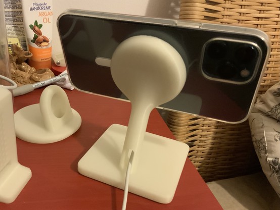 An iPhone mounted in landscape mode on a white plastic stand. The phone is attached with its magnet backside to a disk raised by a short tilted pole from a square base plate. The iPhone stand is made from white plastic with a 3D printer. Left to the iPhone stand are two other 3D models of stands for Apple Watch charger from the same white material.