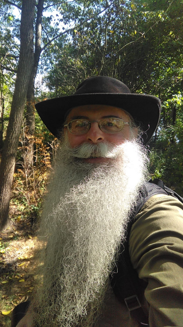 It's a me! 

Adventurous black hat, silver eye glasses,  long nineteen inch white beard with a full moustache.  Olive work shirt.  Background blue sky, trees and mostly bushes .

Very wizardly appearing Caucasian. 

Photo by Douglas @DarkWaterPhotos @DarkWaterPhotoMedia