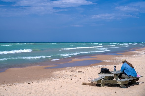 A woman sits at a picnic table on the beach watching the water.