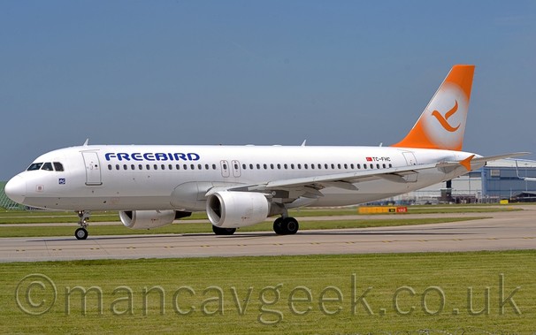 Side view of a twin engoinned jet airliner taxiing from right to left, turning slightly away from the camera.
The plane is mostly white, with a grey belly, and blue "FREEBIRD" titles on the upper forward fuselage.
There are orange vertical endplates on the wingtips.
The tail is orange, with a white circle containing an orange flying bird.
green grass fills most of the foreground, as well as a lare bit of background visible under the planes body.
There is a large grey hangar on the left of the frame, behind the planes tail.
Above, the sky is a pale blue.