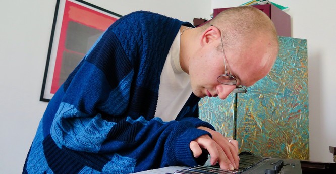 A white man - the composer Laurence Osborn - is hunched intently over a synthesizer keyboard. He has a shaved head and is wearing glasses, a white t-shirt and a knitted cardigan made up of patches which are all different shades of blue. Behind him on the white wall is a print of a painting by Mark Rothko in reds and blacks; behind his left shoulder (he is photographed from his right) is what appears to be a cupboard which has been painted with a splattered sort of textural finish, mostly turquoise, with some gold. The man's hands are close together, with his fingers touching the keys of the keyboard.