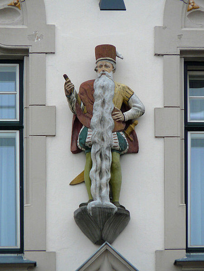 A photograph of a statue on the facade of the Rathaus of Braunau am Inn, Austria.
On a plinth between two windows stands the statue of a man dressed in stockings, pantaloons, tunic and cape, with a cylindrical hat and carrying in his right hand a short ceremonial staff of some kind. A sword is fastened to his belt. Also he has a wide beard which extends down to his feet, and the end is even hanging over the lip of the plinth he's standing on.