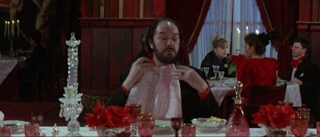 A still from the film The Cook, The Thief, His Wife And Her Lover. A middle aged white man seated at a sumptuously appointed restaurant table tucks the end of a napkin into his collar.