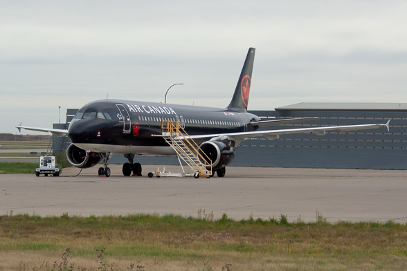 A black jetliner with white under belly facing to the left. There is a set of yellow railed air stairs next to the right engine. A ground power unit hooked up on the left. There is a blast fence behind the airplane. The sky is cloudy gray.