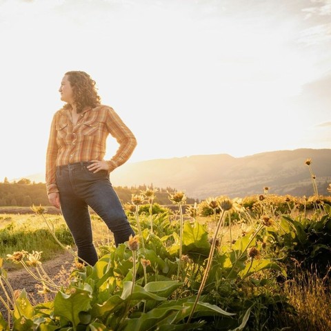 A white woman leans to the left, her hand on her hip. She stares off the left as well. She is standing at the edge of a dirt path, surrounded by yellow flowers. In the background are low mountains. The sun is brilliant behind her, letting off a yellow light. She is wearing blue jeans and an orange and yellow plaid flannel shirt. She has shoulder-length brown curly hair.
