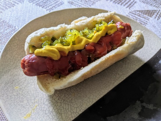 Hotdog in a bun with sweet pickle relish, yellow mustard, and ketchup, on a ceramic plate