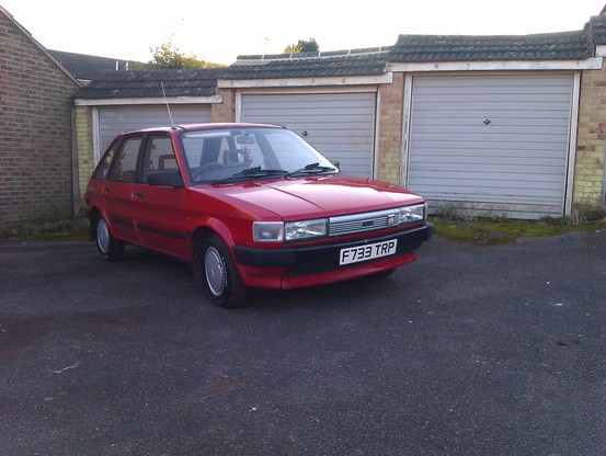 A red 1988 Austin Maestro parked in front of some 1960s Council lock-up style garages with grey up-and-over doors.