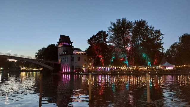 Friedliche Abendstimmung am Wasser. Links im Bild eine geschwungene Brücke, die zu einem Turm auf einer Insel führt. Dort sieht man die bunten Lichter einer Restauration, die sich gemeinsam mit den Gebäuden, den Bäumen und dem Abendhimmel im glitzernden Wasser spiegeln.