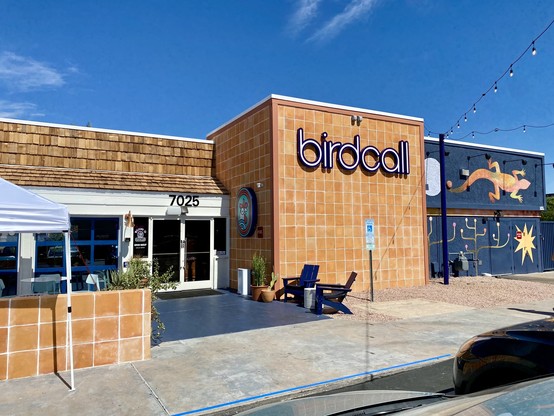 The front entrance of a restaurant on a blue sky day. The restaurant is named Birdcall, and there is gecko artwork with some other stuff on one wall.