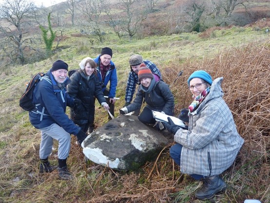 A group of archaeologists wrapped up warm in coats and hats as they inspect a large rock.