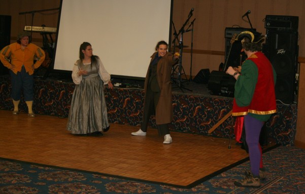 Four actors perform a "Doctor Who" parody play in a hotel conference center ballroom in front of a band stage (with musical instruments) during a science-fiction convention portraying a parody of the 10th Doctor aiming his sonic screwdriver at an someone who (in the play) is an actor portraying the 16th Century commedia dell'arte character of il Capitano while another actor in a silver Renaissance dress watches and the final person is Dottore Who's companion disguised as a 16th Century dressed young man.
