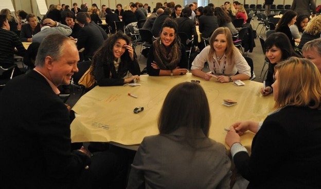 run The Solution Room online: photograph of an in-person Solution Room session. Many round tables of eight people fill a large room.