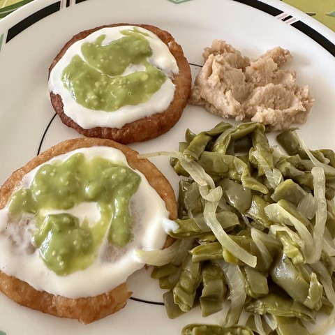 Photo of a plate with two potato patties topped with sour cream and green salsa, with a side of pickled cactus salad and refried Peruvian beans